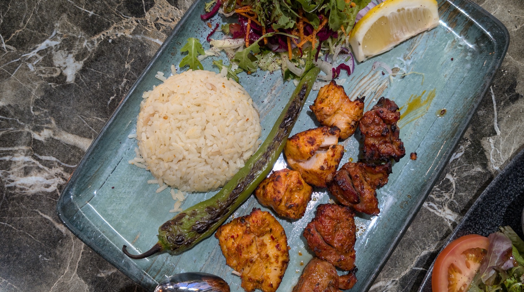 Gegrilde stukken kip met rijst, salade en een gegrilde peper op een langwerpig bord